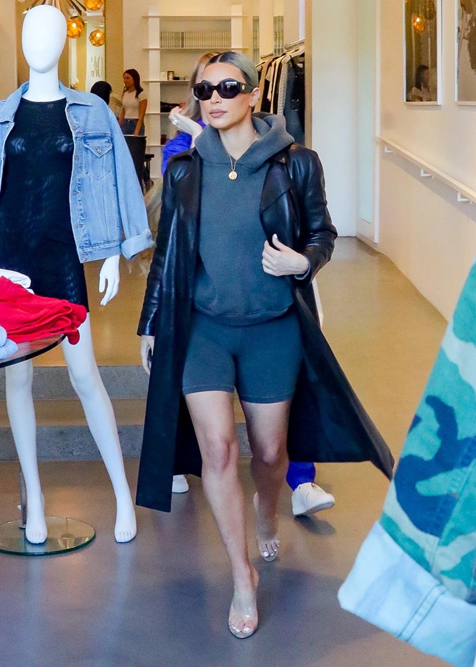 A empresária Kim Kardashian, uma mulher branca de meia idade, com cabelo liso loiro, andando por lojas no shopping. Usa um short de ciclista e um moletom, tudo cinza; um sobretudo preto de couro; uma sandália de salto de plástico transparente. - Metrópoles