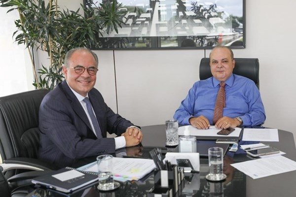 Fotografia colorida mostra dois homens sentados em mesa de reunião