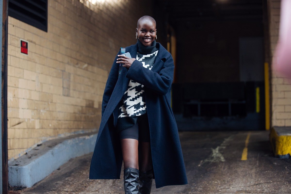 A modelo Adut Akech, uma mulher negra e jovem, de cabelo raspado, posa para foto andando na rua. Ela usa short de ciclista preto, um tricô preto e branco, sobretudo preto e uma bota de cano alta preta de couro. - Metrópoles