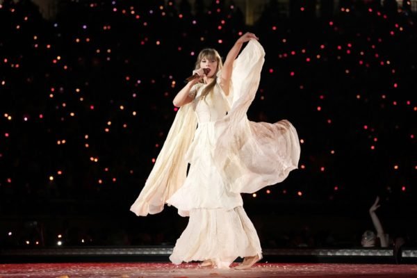 A cantora Taylor Swift, uma mulher jovem e branca, de cabelo loiro liso. Ela está no palco, na turnê The Eras Tour, e usa um vestido branco longo, com camadas e babadas, da marca Alberta Ferretti. - Metrópoles