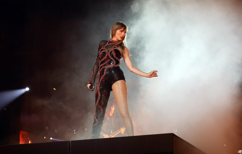 A cantora Taylor Swift, uma mulher jovem e branca, de cabelo loiro liso. Ela está no palco, na turnê The Eras Tour, e usa um macacão preto da marca Roberto Cavalli com uma cobra vermelha bordada. - Metrópoles