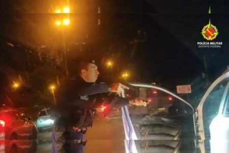 Policial militar fardado abre porta de carro branco e aponta pistola para condutor