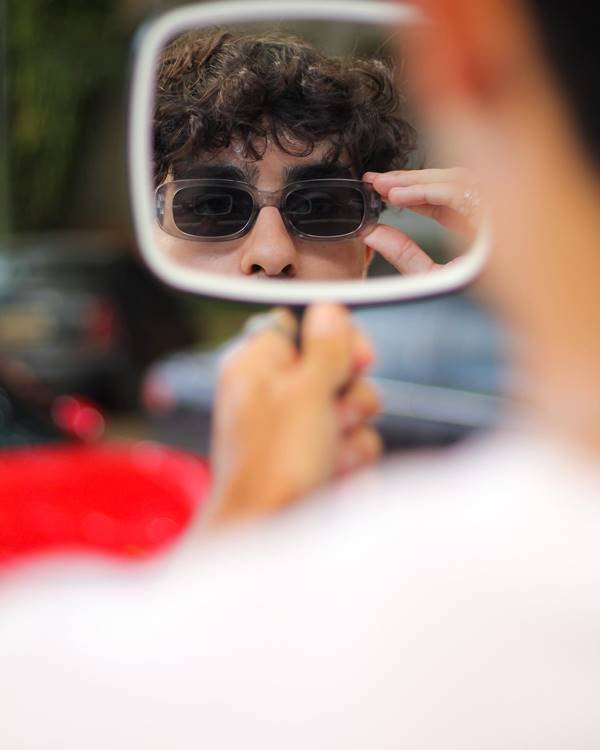 Modelo homem de cabelos ondulados se posiciona em frente a espelho com óculos escuros - Metrópoles 