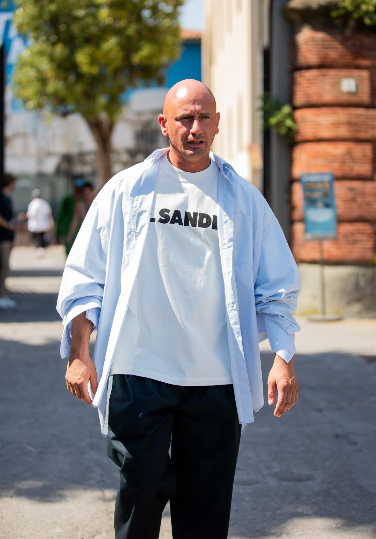 Homem branco e de meia idade, careca, andando pela rua duranta Semana de Moda de Milão. Usa uma camiseta branca, com a palavra Jil Sander escrita, uma calça preta e uma camisa azul de botão. - Metrópoles