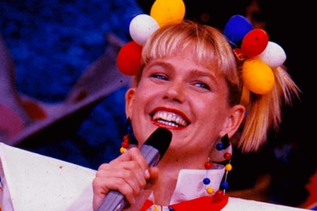 Foto colorida de Xuxa Meneghel. Ela está com bolinhas coloridas na cabeça, batom vermelho, é loira e tem o olho claro - Metrópoles