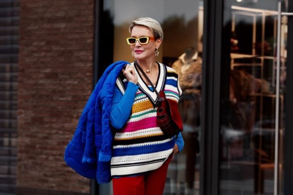 Mulher com colete colorido parada na rua. Ela está segurando uma terceira peça de roupa nos ombros e está usando óculos escuros - Metrópoles