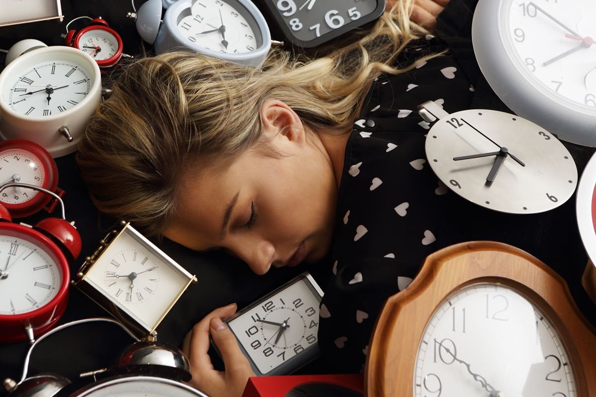 Foto colorida de mulher branca e loira dormindo com vários despertadores ao redor - Metrópoles