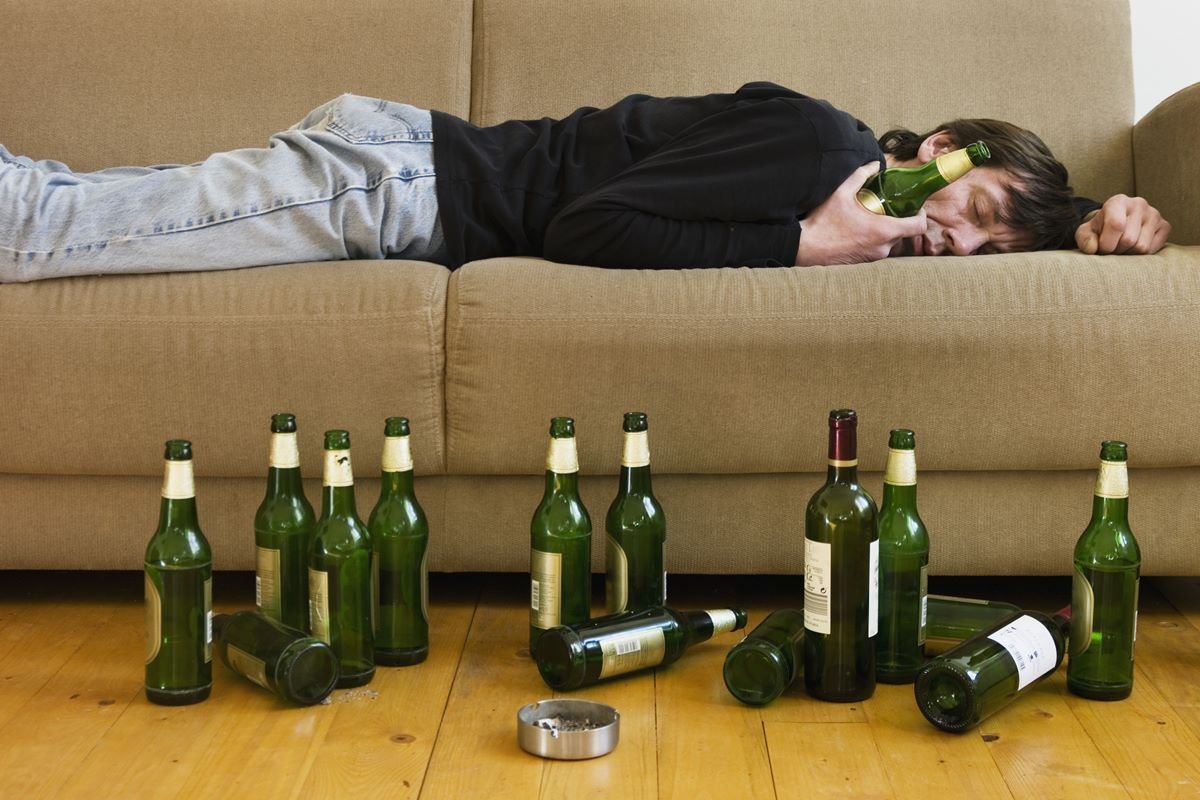 Foto colorida de homem dormindo no sofá segurando uma garrafa de cerveja. No chão, há várias garrafas - Metrópoles 