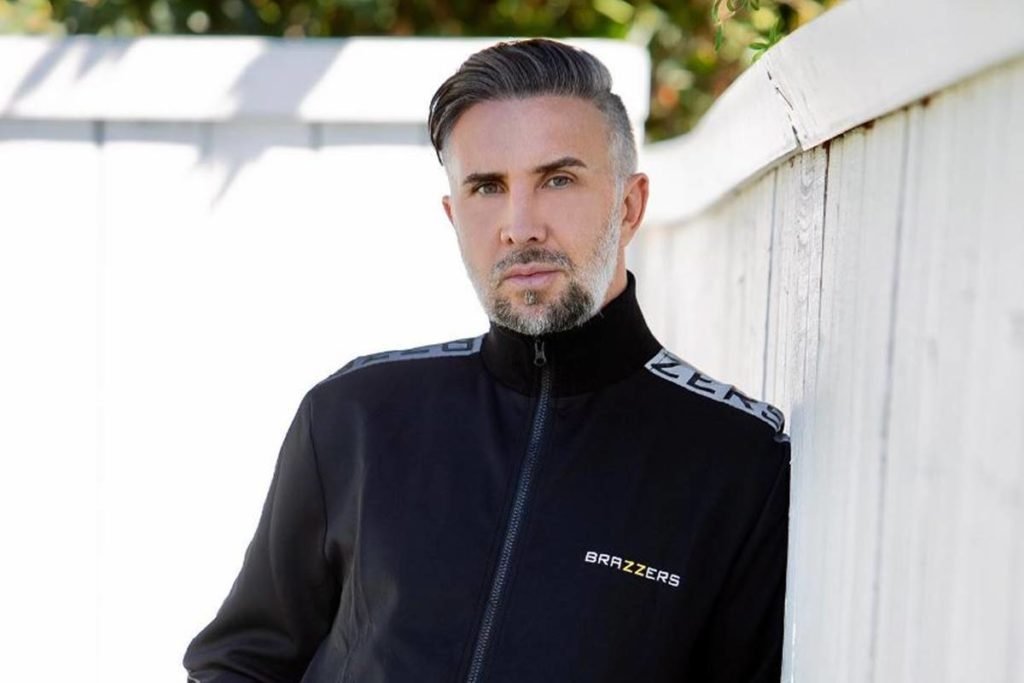 homem branco de cabelos grisalhos usa casaco preto e posa encostado em cerca de madeira - metrópoles