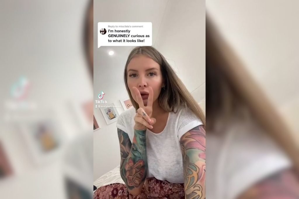 Mulher tatuada vestida de branco segurando celular com uma mão e fazendo sinal de dois com a outra