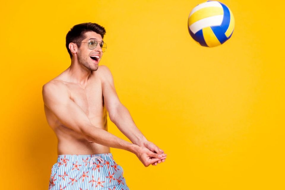Homem sem camisa jogando uma bola de volei para cima e sorrindo - Metrópoles