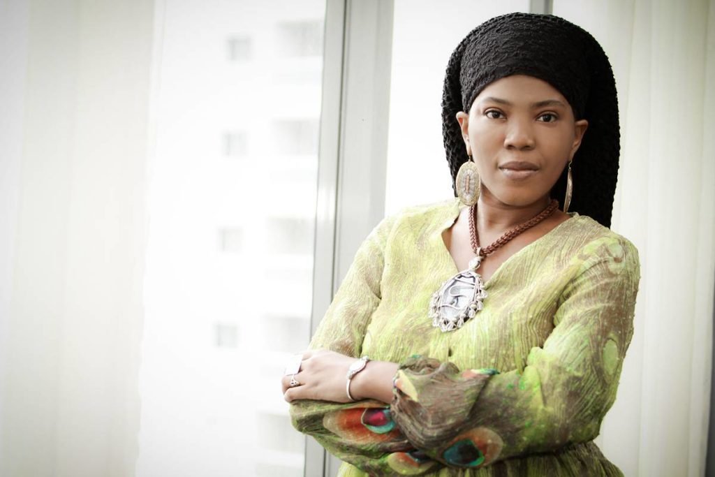 mulher preta posa séria de braços cruzados vestindo um vestido verde mangas longas e um turbante preto - metrópoles