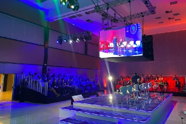 Foto colorida de um auditório com painel de transmissão suspenso - Metrópoles
