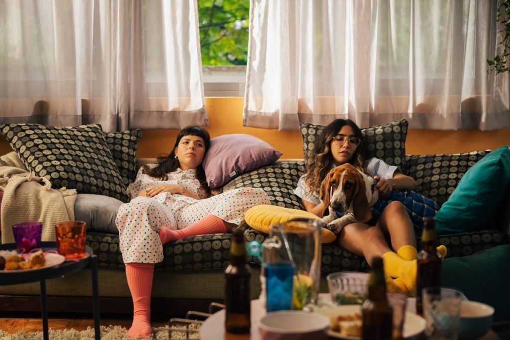 duas jovens sentadas em sofa, uma delas com cachorro no colo - metrópoles
