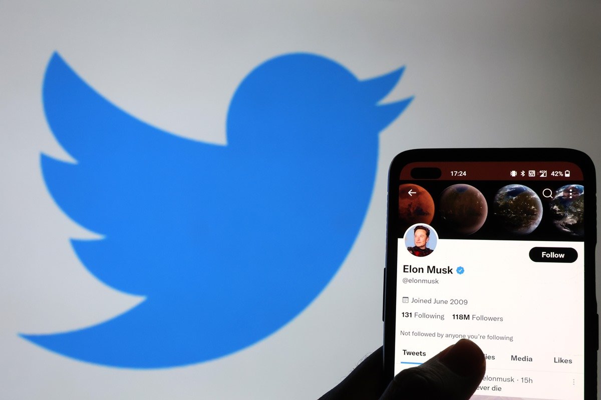 Foto colorida de ícone de passarinho do Twitter ao lado de um celular com a conta do Twitter de Elon Musk - Metrópoles