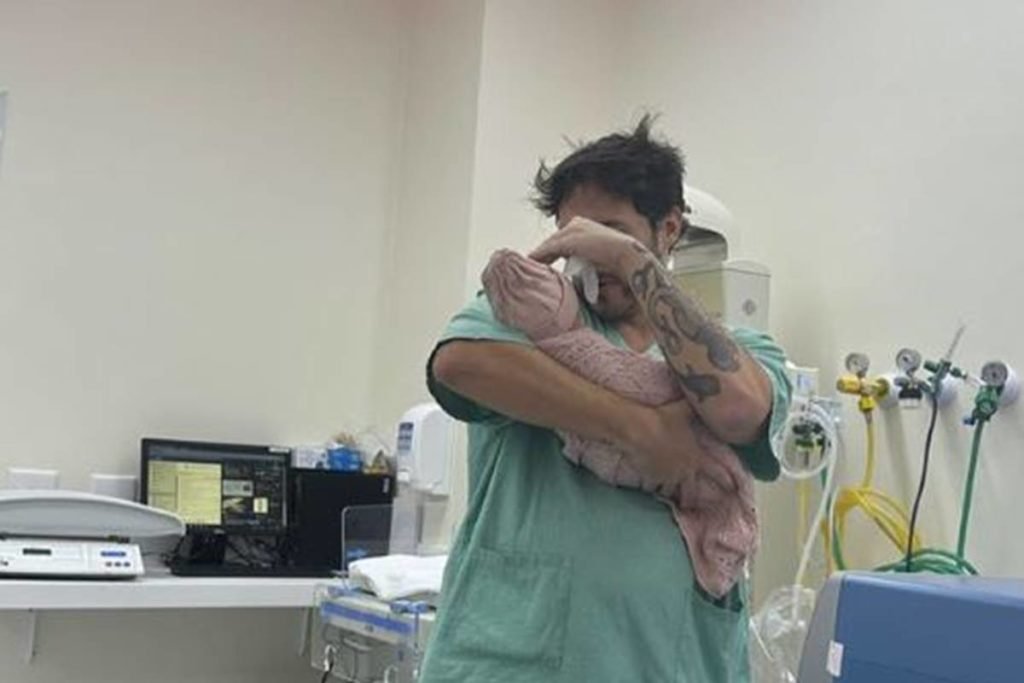 homem de roupa de hospital verde com bebe no colo - metrópoles