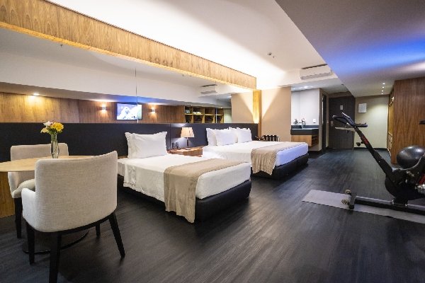 Foto colorida de um quarto com uma mesa e duas poltronas, uma cama de solteiro e uma cama de casal - Metrópoles