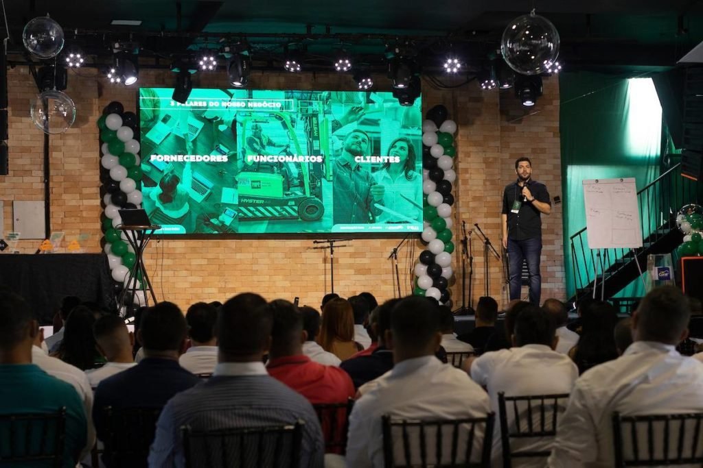 Fotografia colorida mostrando conferência da Ac Coelho com homem falando em cima de palco-Metrópoles