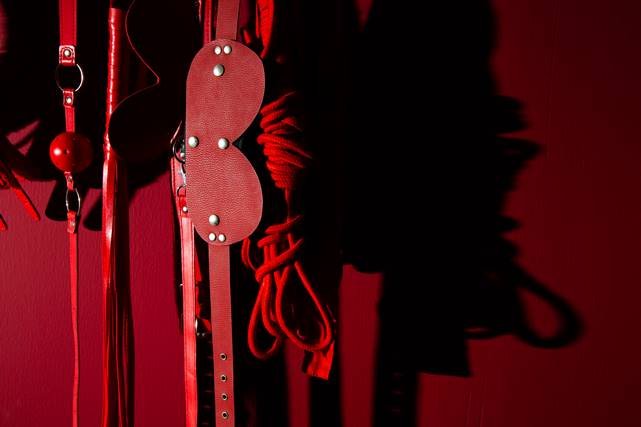 Foto colorida de uma parede vermelha com diversos acessórios fetichistas vermelhos pendurados - Metrópoles