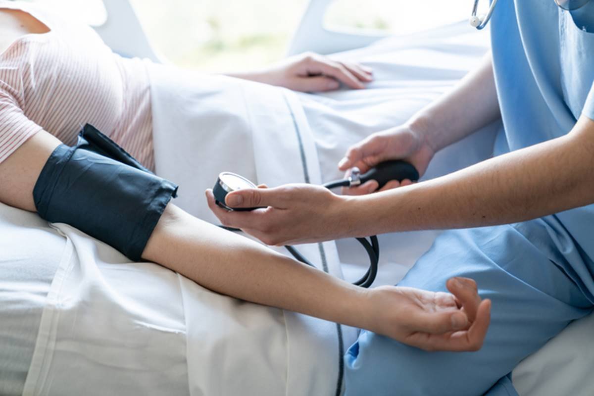 Imagem colorida mostra profissional da saúde vestida de azul medindo pressão arterial de jovem jovem deitada em maca de hospital - Metrópoles