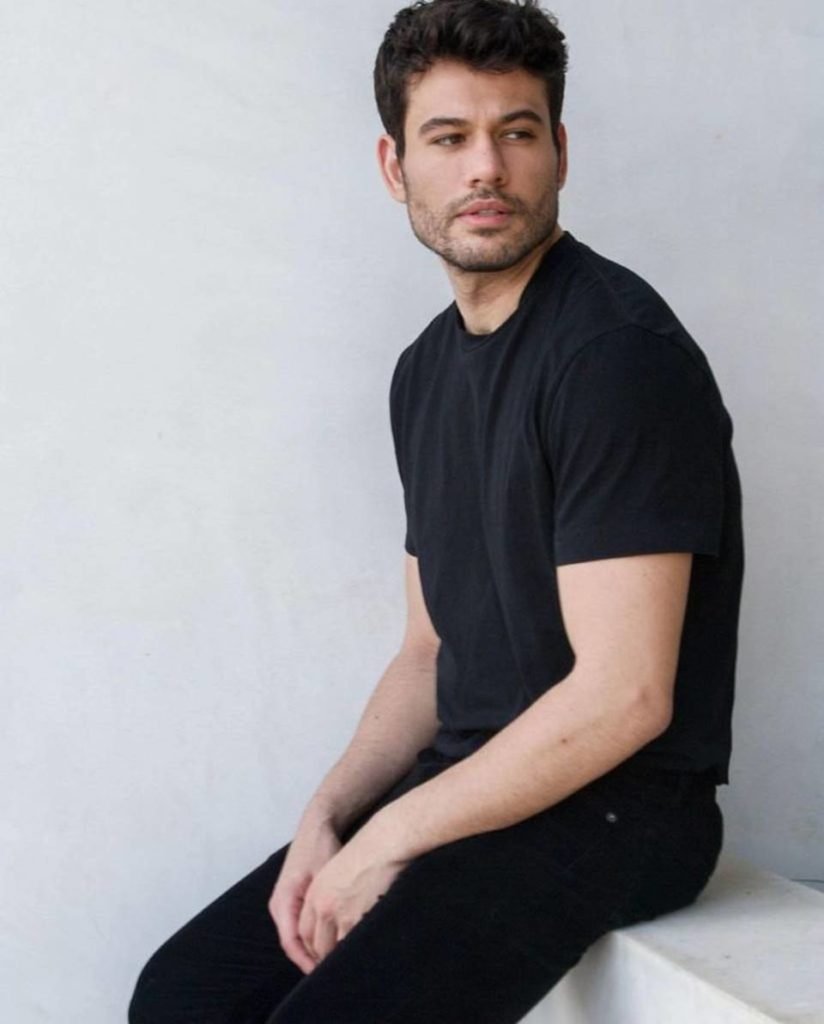 homem de calça e camiseta preta posa sério encostado em mureta branca - metrópoles