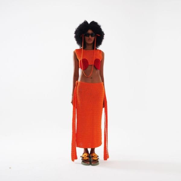 Na imagem com cor, mulher negra com cabelos pretos usando um conjunto cropped e saia laranjas de crochê - Metrópoles