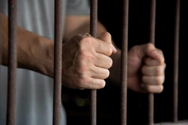 Imagem colorida: mãos de homem dentro de cela de prisão - Metrópoles