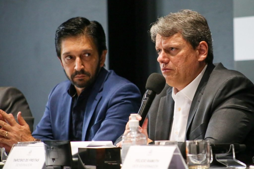 Imagem colorida de Ricardo Nunes (esquerda) e Tarcísio de Freitas (direta). Eles estão sentados e Tarcísio segura um microfone - Metrópoles