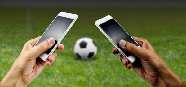 Fotografia colorida mostrando duas mãos mexendo em celulares e uma bola de futebol entre elas-Metrópoles
