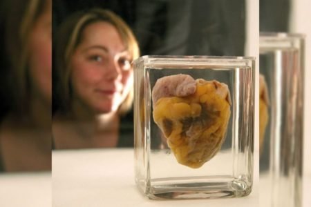 Coração dentro de frasco em conserva focado na imagem, enquanto há uma mulher em plano de fundo desfocada atrás do coração