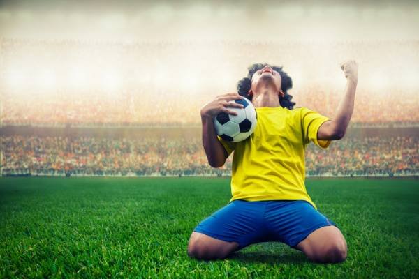 Fotografia colorida mostrando homem com camisa amarela e short azul segurando bola em campo de futebol-Metrópoles