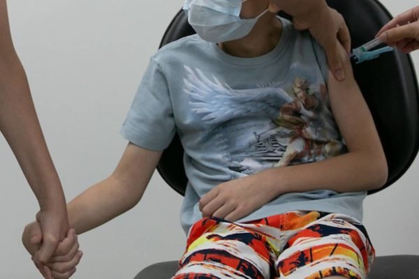 imagem colorida mostra criança branca sentada em cadeira com camiseta azul e bermuda colorida, com máscara no rosto. um adulto segura a mão da criança, que é vacinada com agulha no braço esquerdo