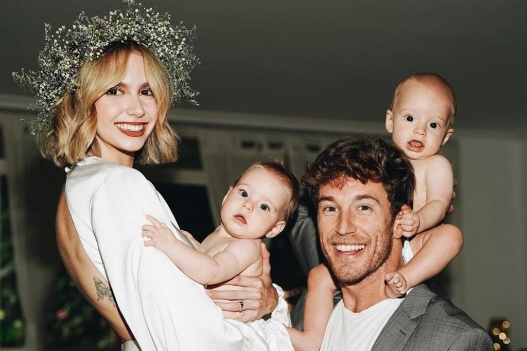 isa scherer vestida de noiva posa com um dos filhos no colo ao lado de rodrigo calazans, de terno cinza, com o outro bebe nos ombros - metrópoles