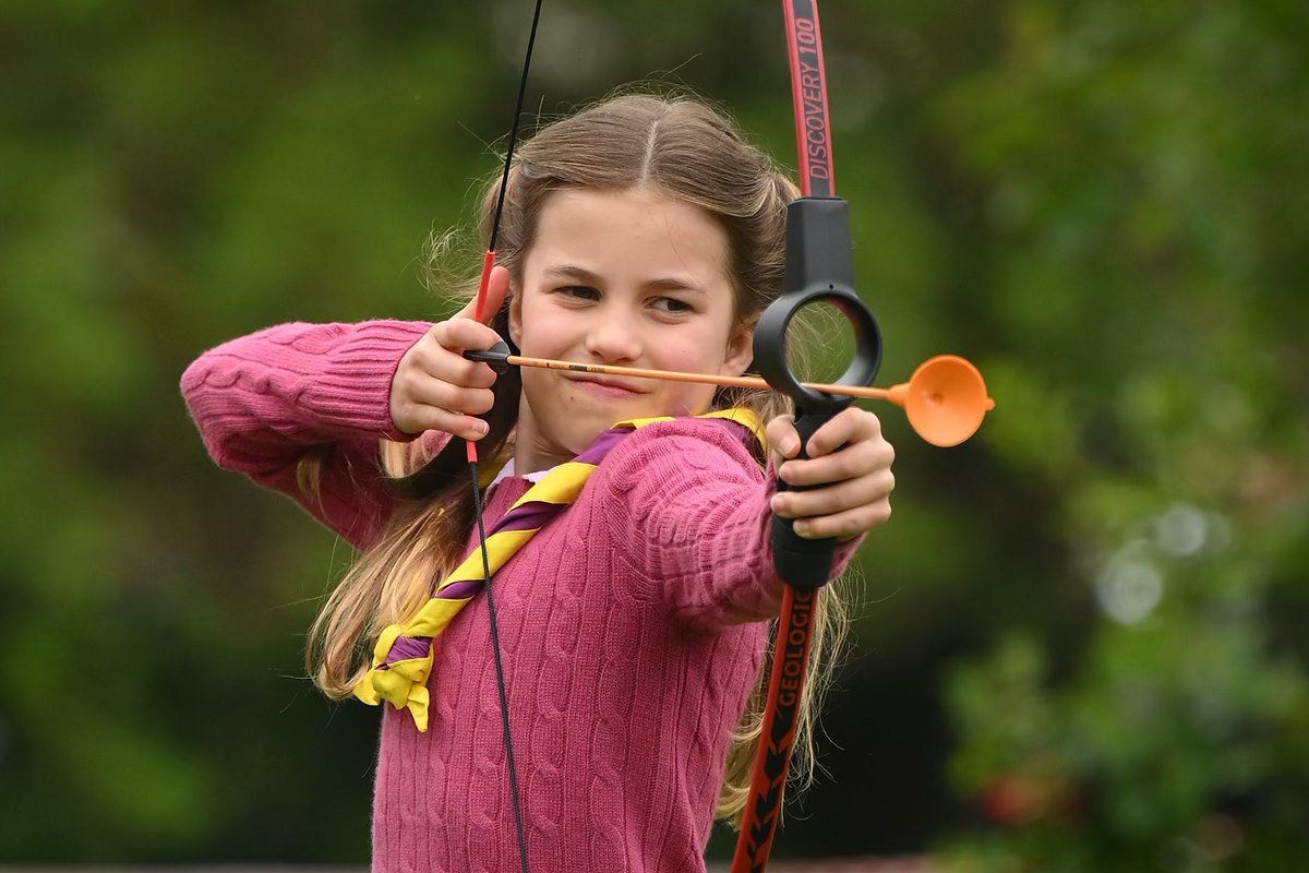 Foto colorida de menina branca e loira atirando com um arco e flecha