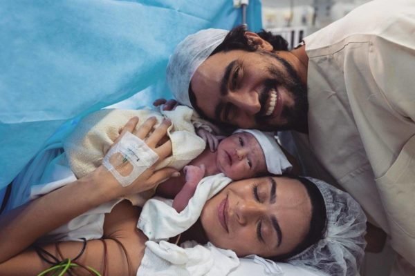 Foto de um homem, um bebê e uma mulher logo após o parto. Eles estão em uma cama de hospital e com roupas apropriadas para o local - Metrópoles