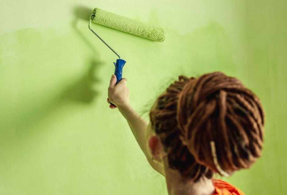 Uma linda designer de mulher faz reparos pinta uma parede cinza com tinta  verde com um pincel camisa xadrez cabelo comprido e jeans reparo no  apartamento