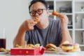 Homem de blusa cinza sentado a mesa comendo frituras como batatas fritas , hamburguer - Metrópoles