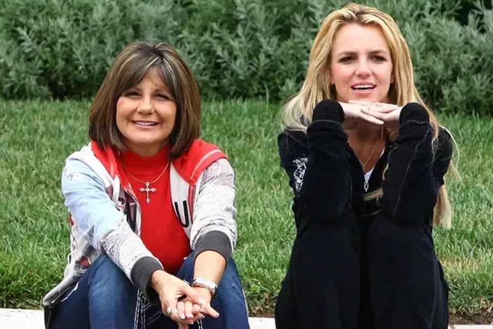 Foto colorida de Britney Spears com a mãe Lynne Spears. A mãe tem cabelo castanho curto, está sorrindo e usa uma blusa vermelha e calça jeans. Britney está com look preto e cabelo loiro. As duas estão sentadas e ao fundo é possível ver um gramado - Metrópoles
