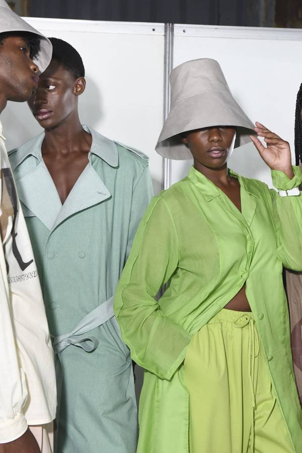 Em backstage de desfile, modelo usa look verde com camisa e chapéu cinza, ao lado de modelo com vestido de alfaiataria com lapela - Metrópoles