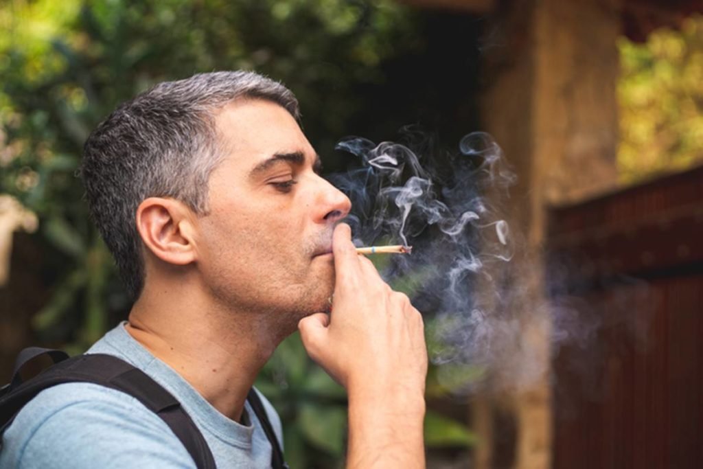 Cigarro de palha: homem com cabelos grisalhos, de mochila, de azul, de perfil fumando cigarro