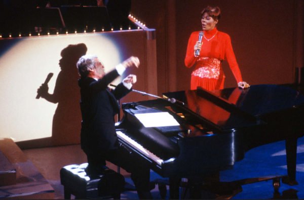 Na imagem com cor, uma mulher negra cantando ao lado de um piano usando vestido vermelho - Metrópoles