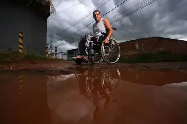 Fotografia colorida mostra homem de cadeira de rodas em rua com lama
