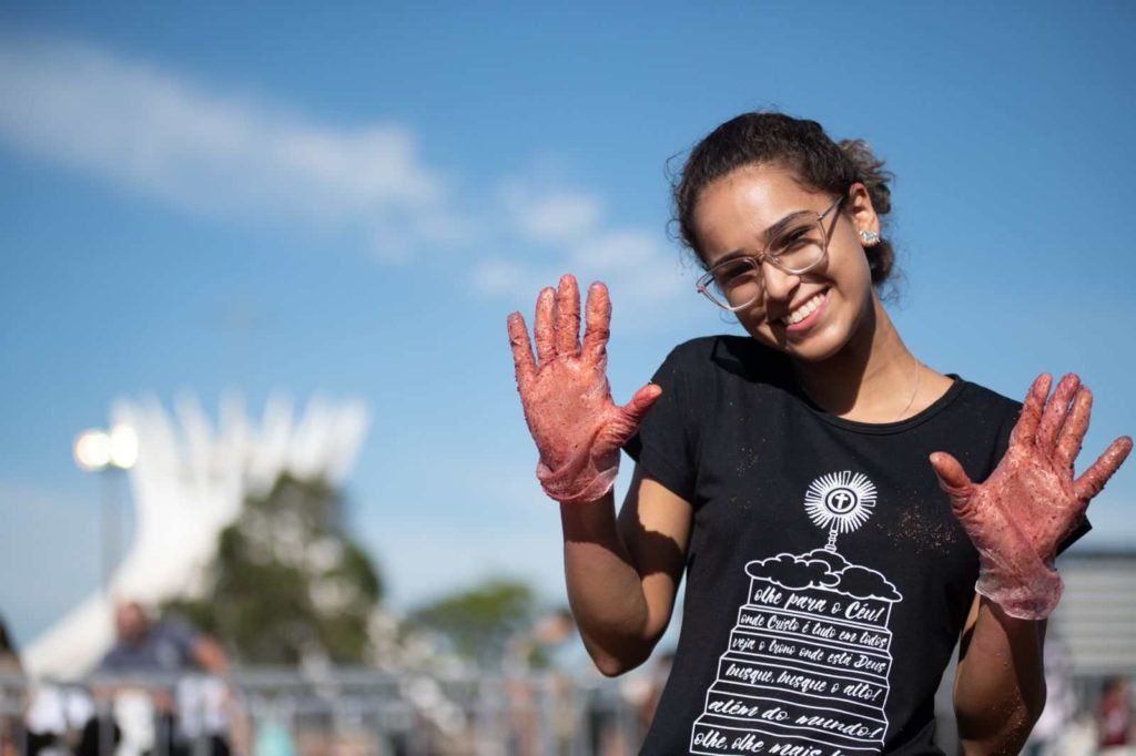 Mulher de cabelos cacheados mostra luvas cheias de terra. Ela veste camisa azul escura. Catedral de Brasília ao fundo