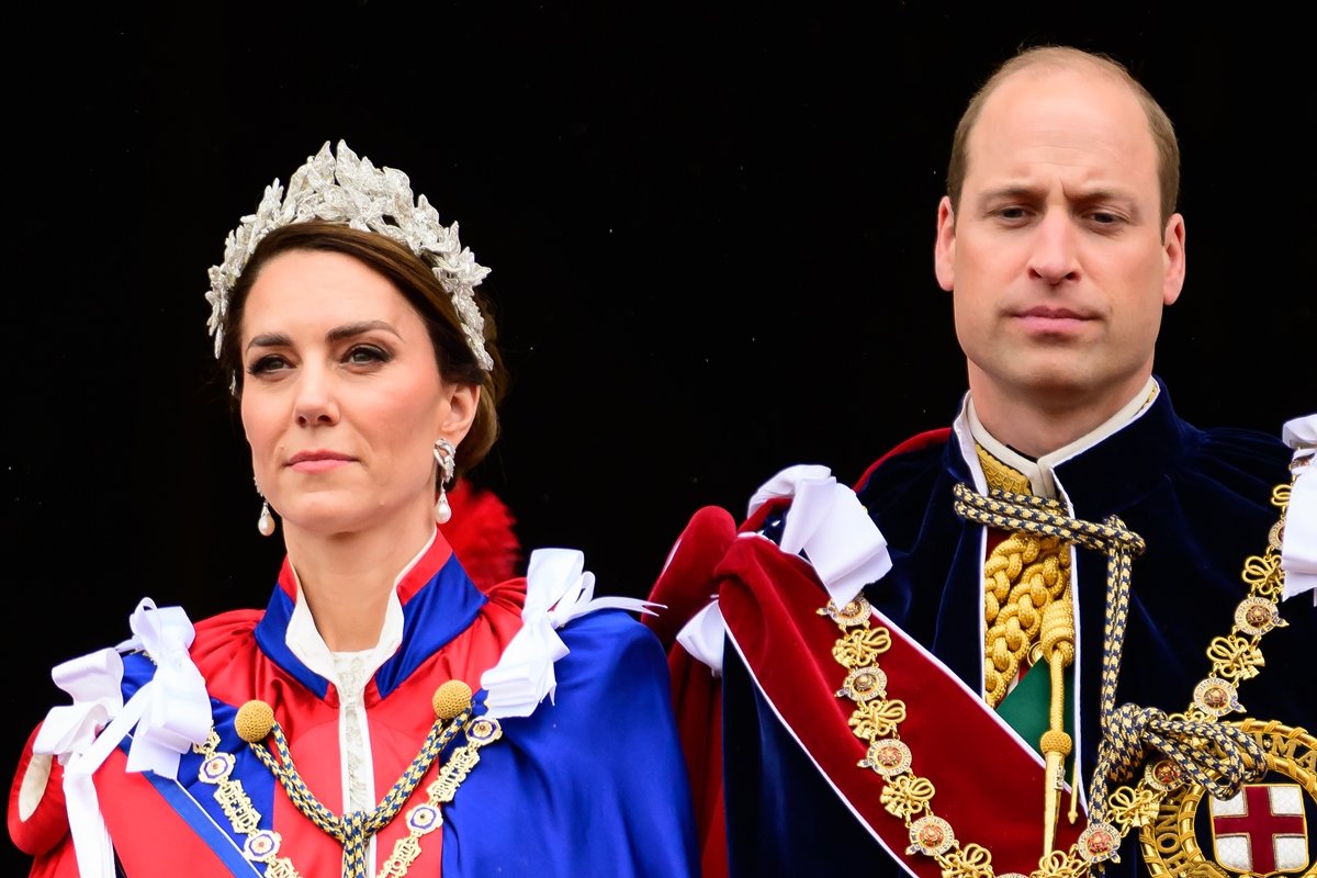 Kate está usando uma tiara prara e uma capa azul com vermelha e detalhes brancos. WIlliam está olhando seriamente para o lado esquerdo. Ele está vestido uma capa preta, com uma faix vermelha e detalhes dourados