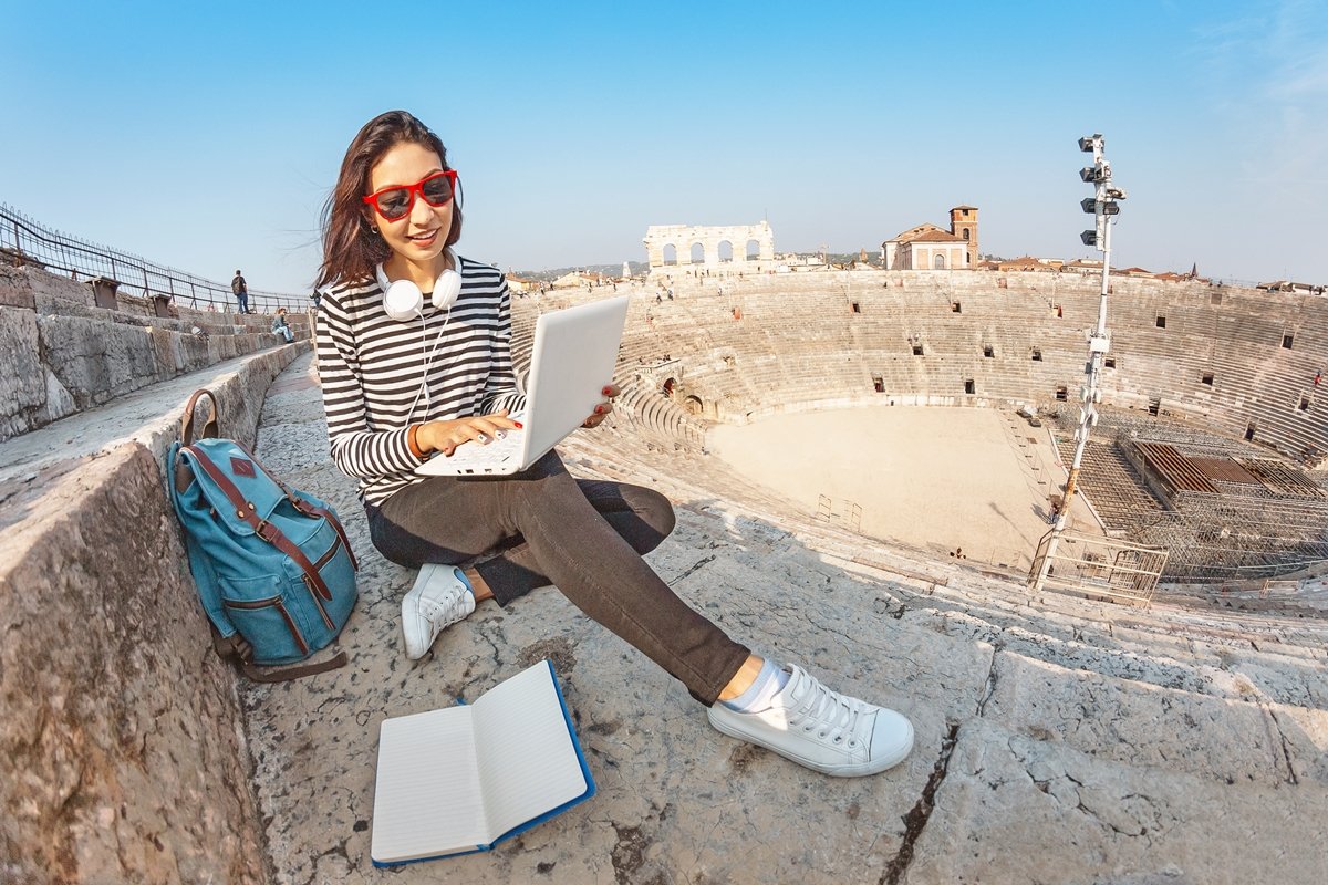 Happy asian girl student doing her homework with notebook pc or freelance working, while sitting in the european landmark amphitheater. Vacation and studying concept