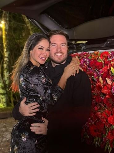Foto colorida de Maíra Cardi e Thiago Nigro. Eles estão abraçados e mostram um carro cheio de flores - Metrópoles