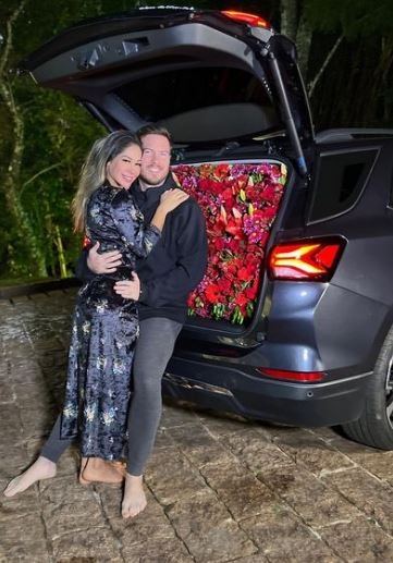Foto colorida de Maíra Cardi e Thiago Nigro. Eles estão abraçados e mostram um carro cheio de flores - Metrópoles