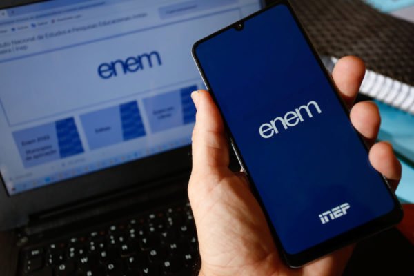 Enem Imagem colorida mostra celular com a tela em uma aplicativo do Enem sendo segurado por uma mão, com um computador ao fundo - Metrópoles
