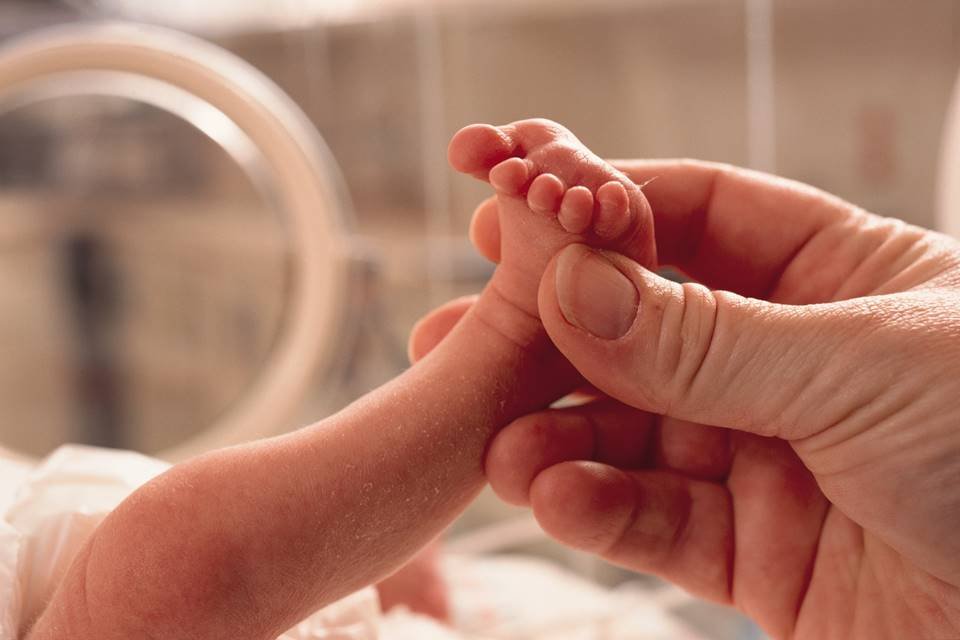 Foto colorida de uma mãe segurando o pé de um bebê dentro do hospital - Metrópoles