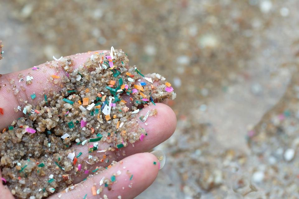Fotografia colorida mostra mão cheia de areia com microplásticos coloridos de plástico - Metrópoles